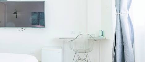 Une chambre d’hôtel minimaliste comprenant un lit, un petit réfrigérateur blanc, une table en verre avec une chaise, un téléviseur à écran plat et un rideau.