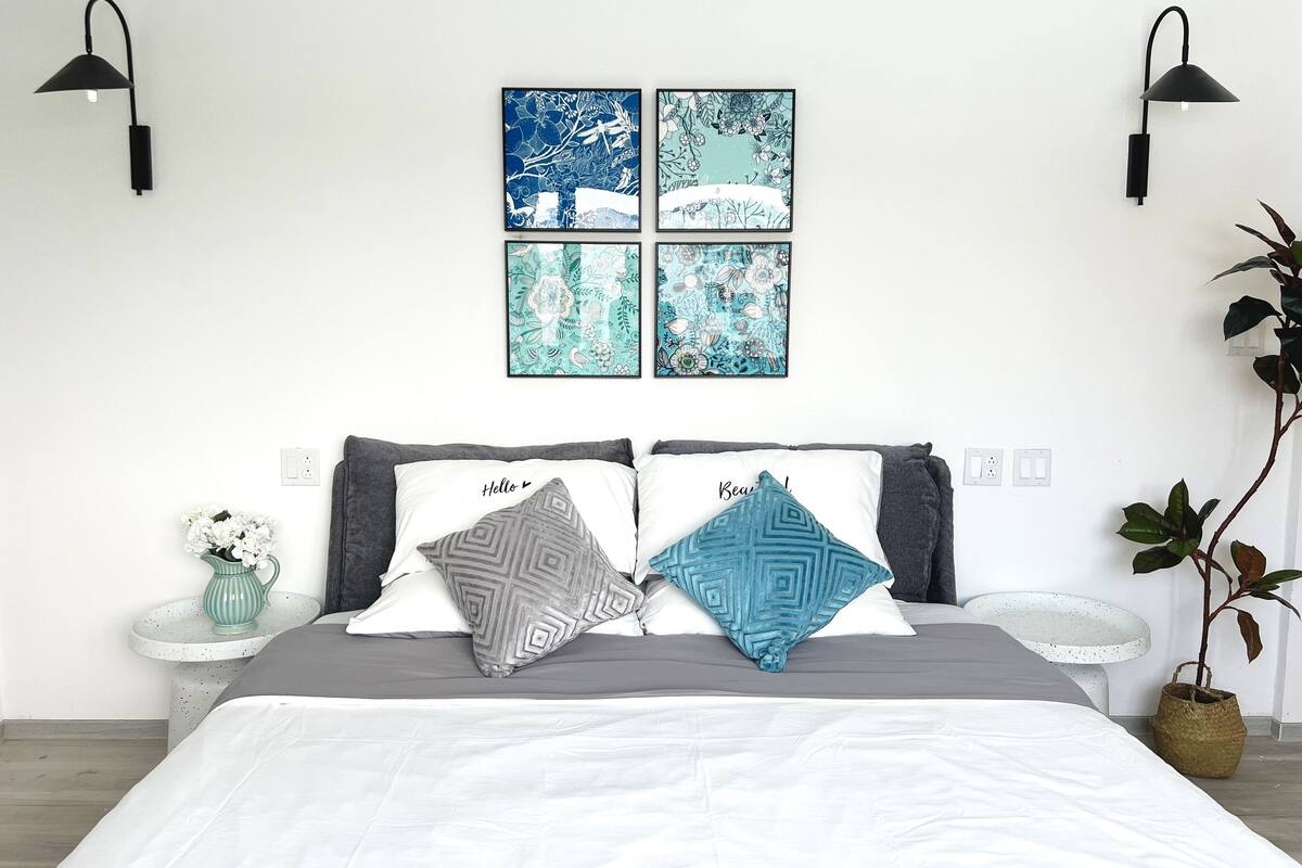 A neatly arranged bedroom with a bed, bedside tables, a wall-mounted lamp, a potted plant, and floral artwork.