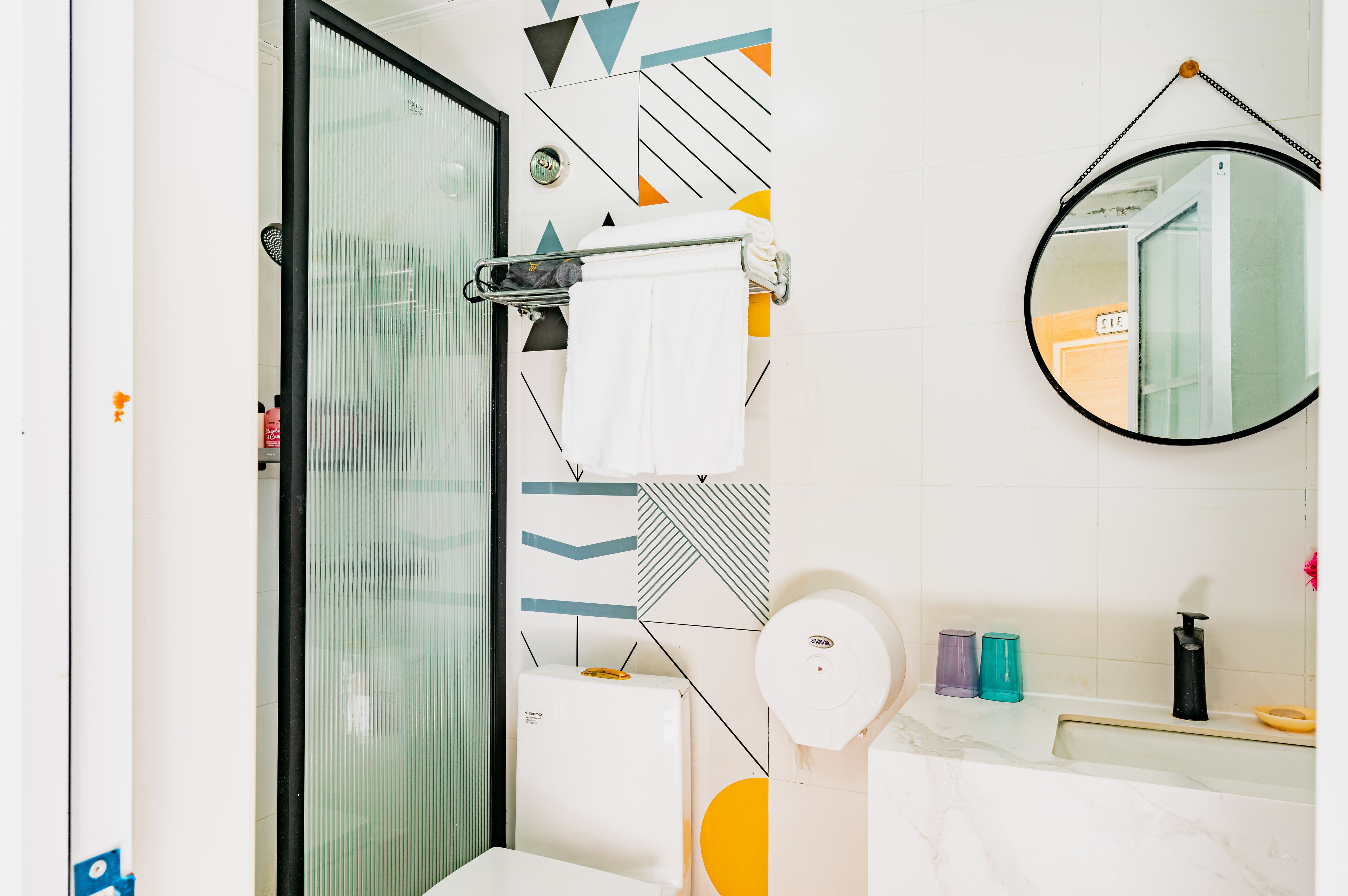 Une salle de bain avec une douche à parois de verre, un porte-serviettes avec une serviette blanche, un miroir rond et un lavabo avec des articles de toilette.
