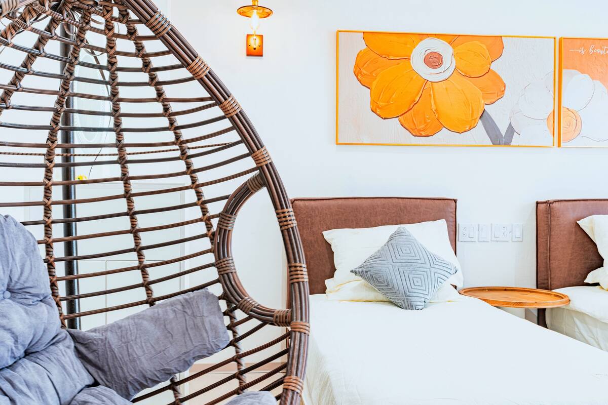 A room with a rattan chair, a bed with white bedding, a grey pillow, and a wall with a flower painting.