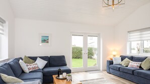 A spacious living room with two dark blue sofas, a wooden coffee table, and a large sliding glass door leading to a garden. - Llwyn Eithin (Holyhead)