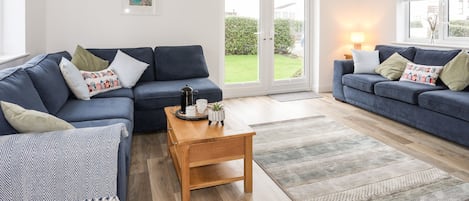 A spacious living room with two dark blue sofas, a wooden coffee table, and a large sliding glass door leading to a garden.
