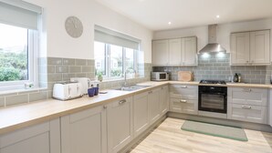 A modern kitchen with grey cabinets, a stainless steel sink, a microwave, and a stove. There are two windows providing natural light. - Llwyn Eithin (Holyhead)