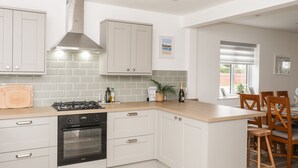 A modern kitchen with white cabinets, a gas stove, an oven, and a large island with bar stools. - Llwyn Eithin (Holyhead)