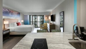 A hotel room with a bed, a desk, a chair, a TV, a bathroom with a sink, and a view of the outdoors. - Beach Getaway Resort View Deluxe King Room 2nd Flr (Myrtle Beach)