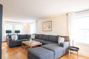 A modern living room with a sectional sofa, a coffee table, and a lamp. - Heated Plunge Pool, Game Barn, AC, 5 Min to Beach! (Chatham)