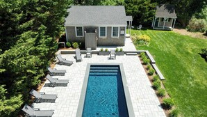 Une piscine rectangulaire entourée d’une terrasse dallée, elle-même bordée d’une pelouse bien entretenue et d’une petite maison dotée d’un porche.