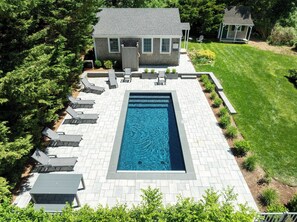 A rectangular swimming pool with a paved deck, surrounded by a well-maintained lawn and a small house with a porch. - Heated Plunge Pool, Game Barn, AC, 5 Min to Beach! (Chatham)