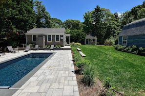 A swimming pool with a stone deck, a small building with a door, and a grassy yard with trees. - Heated Plunge Pool, Game Barn, AC, 5 Min to Beach! (Chatham)