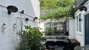 A small outdoor patio with a white brick wall, a large window, and a grill. - Stylish 2 Bedroom, 2 Bathroom Terraced Townhouse & Garden (LONDON)