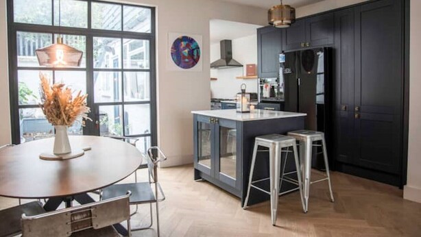 A modern kitchen with dark cabinets, a central island, and a large window. - Stylish 2 Bedroom, 2 Bathroom Terraced Townhouse & Garden (LONDON)