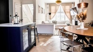 A modern living room with a sofa, a dining table with chairs, a kitchen area with a fridge, and a staircase leading to another floor. - Stylish 2 Bedroom, 2 Bathroom Terraced Townhouse & Garden (LONDON)