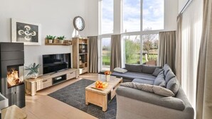 A modern living room with a fireplace, TV, sofa, and large windows offering a view of the outdoors. - Villa Am Gutshof (Trent)