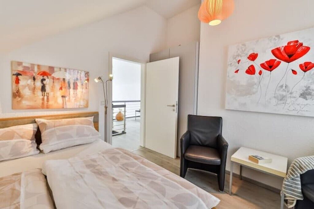 A hotel room with a bed, a chair, a table, and two paintings on the walls.