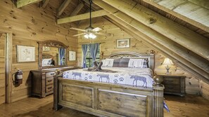 Un dormitorio con piso de madera, una cama, un aparador, una mesita de noche, un ventilador de techo y cuadros enmarcados en las paredes.