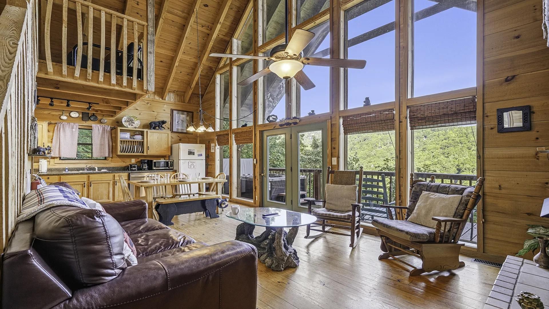 A spacious cabin interior with a dining area, a living room with a leather couch, and large windows offering a view of the outdoors.