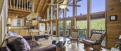 A spacious cabin interior with a dining area, a living room with a leather couch, and large windows offering a view of the outdoors.