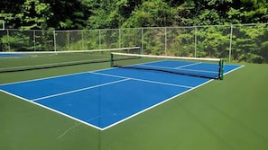 Een tennisbaan buiten met een net, omringd door een hek en groen.