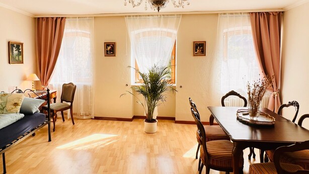 Un comedor con candelabro, mobiliario de madera y grandes ventanales con cortinas.