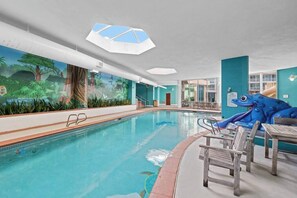 Ein Hallenbad mit einer Dschungel-Muralmalerei, einer blauen Froschskulptur und einer Sitzecke mit Holztischen und -stühlen.