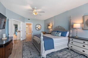 Ein Schlafzimmer mit Bett, Kommode, Nachttisch, Deckenventilator und einer Tür zu einem weiteren Raum.