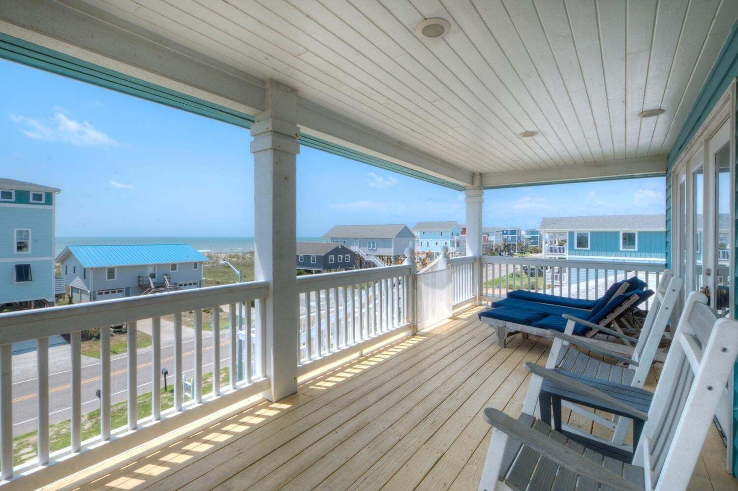 A covered porch with a view of the beach and ocean, wooden deck, and lounge chairs.