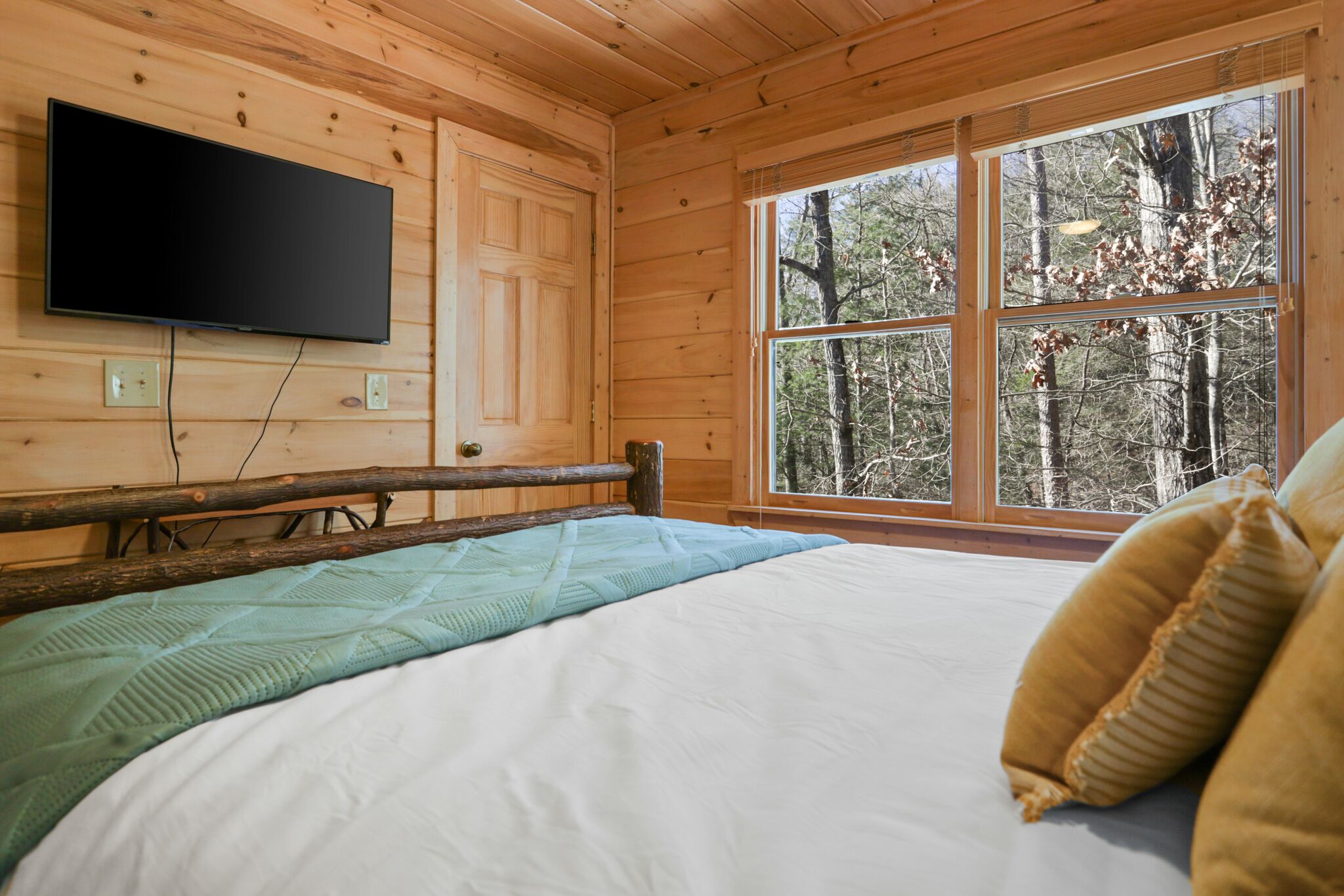 Habitació d'una cabana de fusta amb un llit, televisió penjada i una finestra gran amb vista al bosc.