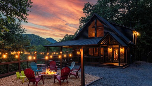 Une cabane dotée d’un foyer extérieur et de sièges, entourée d’une terrasse et illuminée de guirlandes lumineuses.