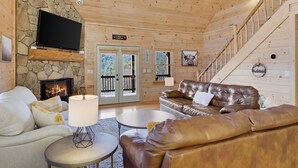L’intérieur d’une cabane en rondins, avec une cheminée en pierre, des canapés en cuir, un téléviseur à écran plat et un escalier.