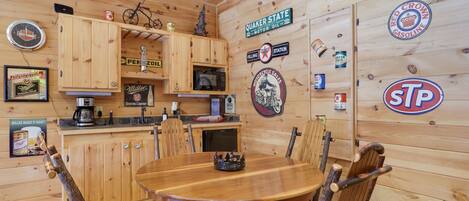 A wooden cabin interior with a dining table, chairs, a coffee maker, and various wall decorations including signs and a clock.
