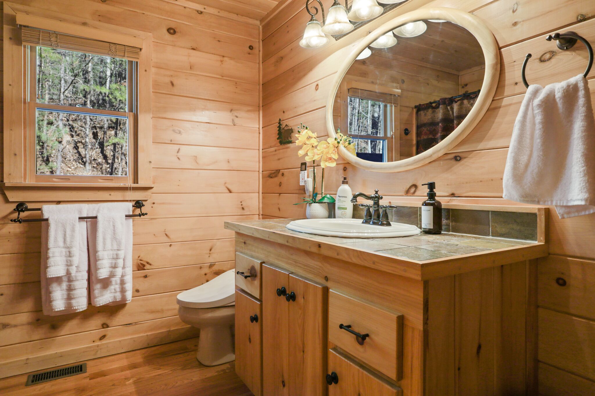 Een houten badkamer in een chalet, met een raam, toilet, wastafel met spiegel en een wasbak.