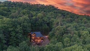 Un chalet à deux étages, entouré d’une forêt dense, doté d’une terrasse visible et d’un éclairage extérieur.
