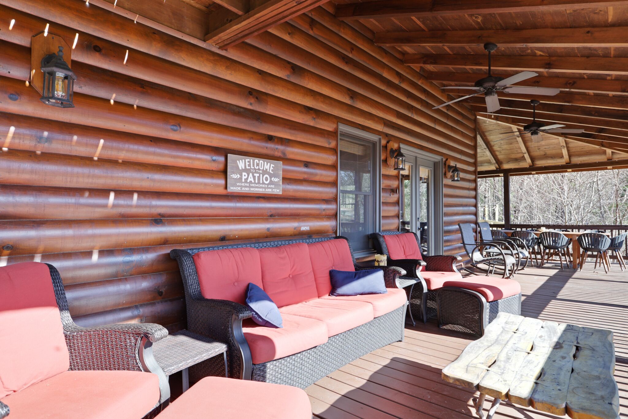 A covered patio with a log cabin exterior, featuring a red cushioned sofa, wicker chairs, and a wooden table.