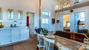 Een ruime keuken met een centraal eiland, een eethoek met een houten tafel en witte stoelen, en een slaaploft met een stapelbed.