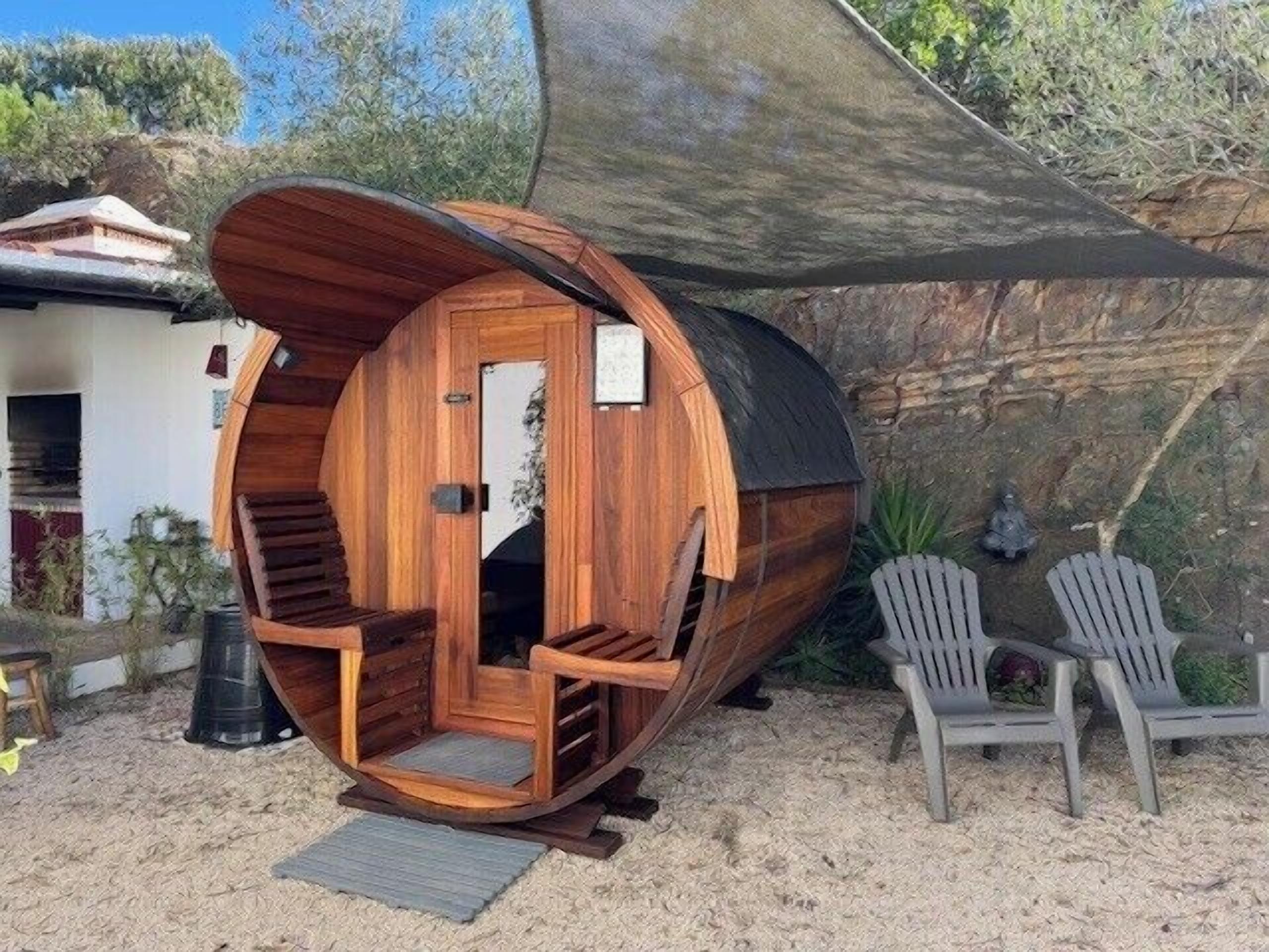 Eine zylindrische Holzkonstruktion mit Tür und Stufen, vermutlich eine Sauna oder ein Whirlpool, umgeben von Sitzgelegenheiten und einem schattigen Bereich.