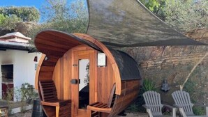 Een houten, cilindervormige constructie met een deur en treden, waarschijnlijk een sauna of hottub, omringd door buitenmeubels en een beschaduwd gebied.
