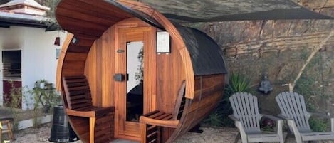 A wooden cylindrical structure with a door and steps, likely a sauna or hot tub, surrounded by outdoor seating and a shaded area.