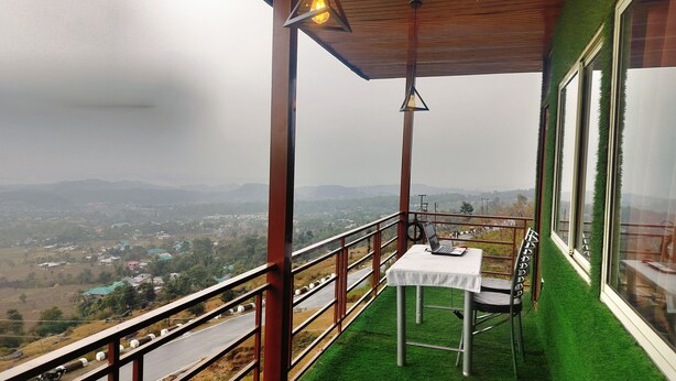 A balcony with a table and chair, overlooking a valley with houses and a road. - Hotel Nirbana Heights (Dharamshala)
