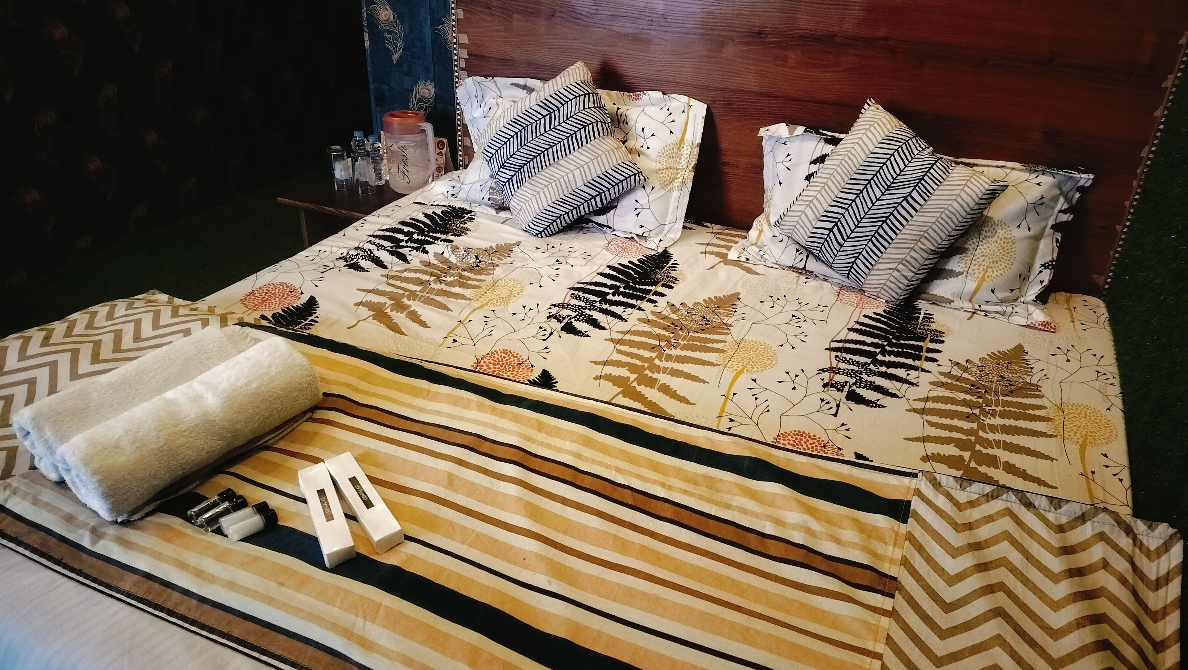 A neatly made bed with patterned bedding and pillows, a rolled-up towel, and a bedside table with a water bottle and salt shaker.