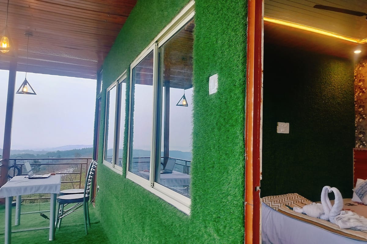 Una habitación con cama, una mesa, sillas, un ventanal con vistas a las colinas y una pared verde.
