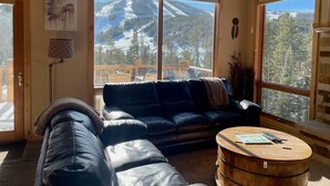 A cozy living room with a large window offering a mountain view, a black leather sofa, and a wooden coffee table. - Antler Way At Lakota.... 3 Bedroom Townhouse by RedAwning (Winter Park)
