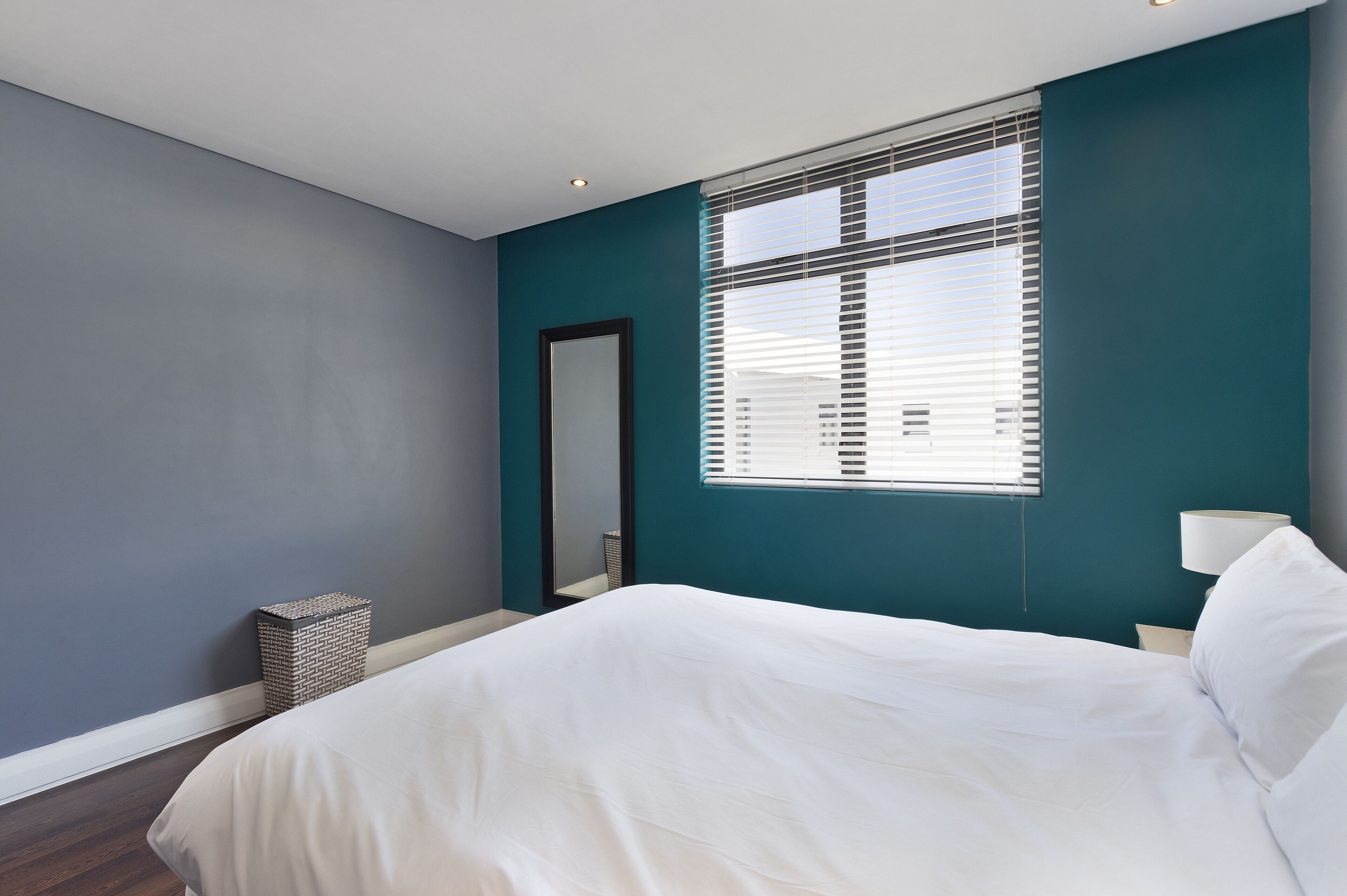 A modern bedroom with a large bed, a window with blinds, a small side table, and a trash bin.