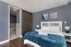 A bedroom with a bed, bedside tables, a nightstand, a wardrobe, and a door leading to a bathroom. - Eden on the Bay 240 (Cape Town)