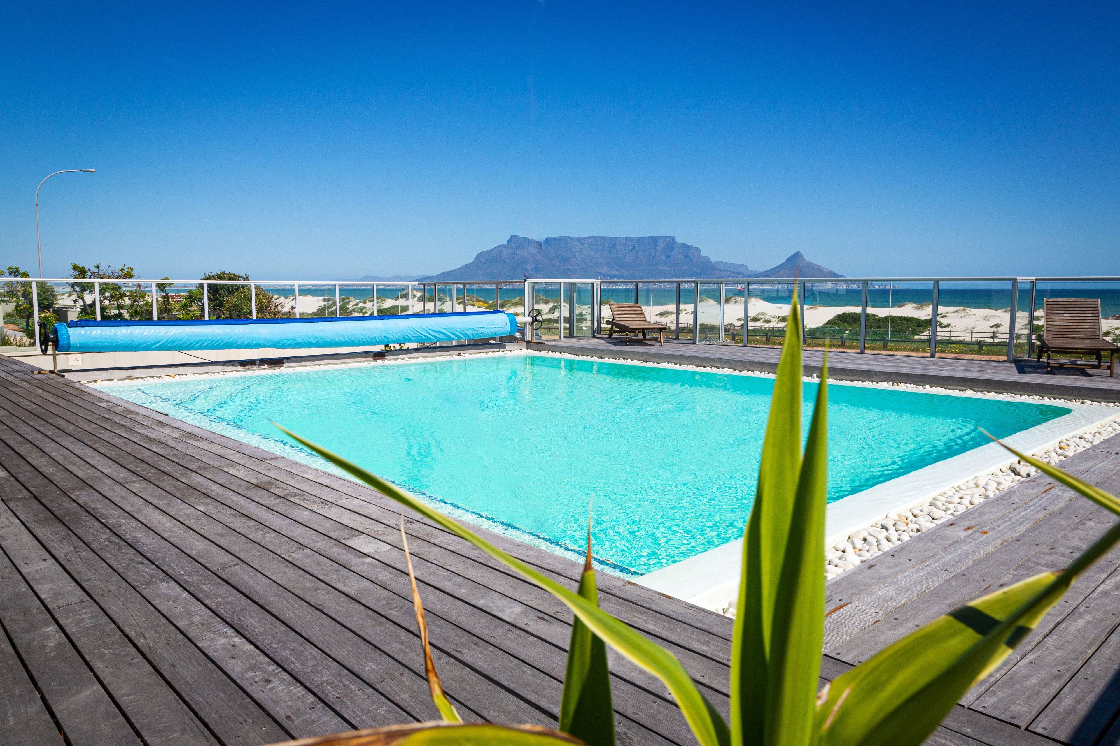 A pool with a wooden deck, lounge chairs, and a view of the beach and mountains.