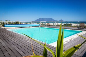 A pool with a wooden deck, lounge chairs, and a view of the beach and mountains. - Sea Spray A308 (Cape Town)