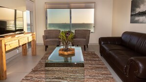Un salon doté d’une grande fenêtre, d’un canapé en cuir marron, de deux fauteuils, d’une table basse en verre ornée d’un vase et d’un meuble TV en bois.