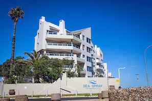 Un immeuble moderne à plusieurs étages, doté de balcons, d’un palmier et d’une enseigne sur laquelle on peut lire « SEA SPRAY ».