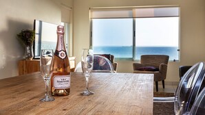 Une table en bois sur laquelle sont posés une bouteille de champagne rosé Simonsig Brut, deux verres, et d’où l’on peut admirer l’océan à travers de grandes fenêtres.