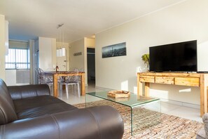 Una sala de estar moderna con un sofá de cuero, una mesa de centro de vidrio, un televisor de pantalla plana sobre un mueble de madera, y un comedor con una mesa y sillas de vidrio.