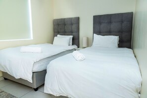 A hotel room with two beds, each with a grey upholstered headboard, white bedding, and a rolled towel on one bed. - Sea Spray A308 (Cape Town)